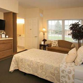 A cozy bedroom with a bed, couch, and television, featuring soft lighting and a large window.