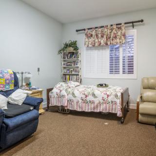 A cozy bedroom featuring a single bed, a blue armchair, and a beige recliner with a bookshelf filled with books.