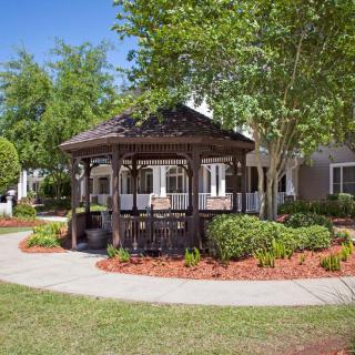 A charming wooden gazebo surrounded by well-maintained landscaping and a paved pathway under a clear blue sky.