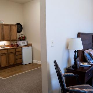A cozy apartment interior showing a small kitchen area and a bedroom with a lamp and chairs.