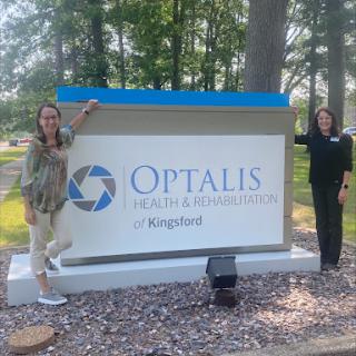 Two women pose beside the Optalis Health and Rehabilitation sign in a park-like setting.