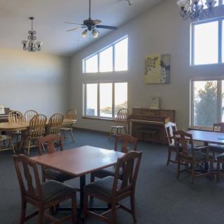 A spacious dining area with large windows, featuring multiple tables and chairs, illuminated by natural light and ceiling fixtures.
