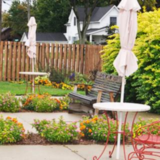 A quaint outdoor patio area with tables, chairs, and closed umbrellas surrounded by a garden and wooden fence.