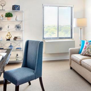 A bright and modern office space with a blue chair, beige sofa, decorative shelves, and a large window.
