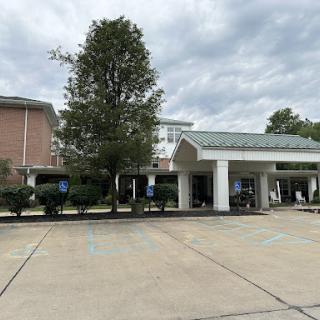 A view of a hotel entrance with a large tree and a parking area under a cloudy sky.
