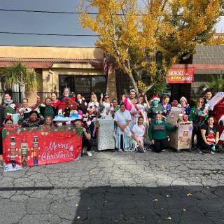 A festive group gathers outdoors for a Christmas celebration, displaying a colorful banner and wearing holiday-themed costumes.