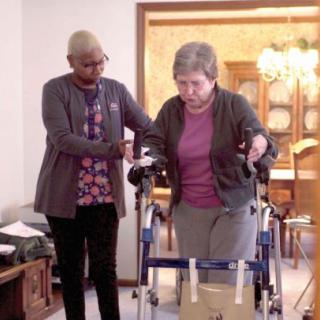 A caregiver assists an elderly woman using a walker in a home setting.