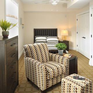A cozy bedroom featuring a patterned armchair, bed, dresser, and decor elements like a lamp and plant.