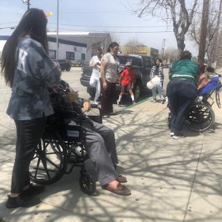 A group of individuals gathered outside, some in wheelchairs, waiting in a community space.