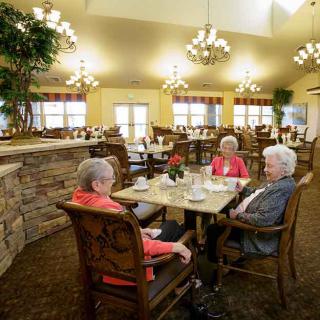 Three elderly women enjoy a conversation in a cozy dining area with chandelier lighting.