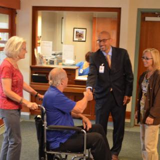 A healthcare professional greets a patient in a wheelchair, while family members look on in a hospital setting.