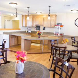 A spacious kitchen and dining area with wooden flooring, featuring several tables and chairs, pendant lights, and decorative flowers.