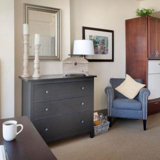 A cozy living room corner featuring a blue armchair, a dark dresser with decorative items, and a warm aesthetic.
