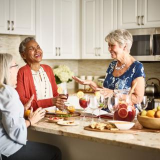A group of people enjoying conversation and laughter in a kitchen while preparing food and drinks.