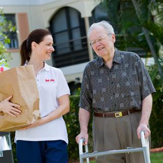 A caregiver assists an elderly man with a walking aid while carrying a bag of supplies.