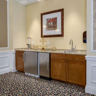 A small kitchenette with wooden cabinets, a sink, and a mini fridge, decorated with framed artwork above the countertop.