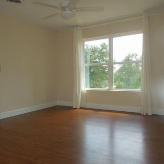 A bright, empty room with hardwood flooring, a large window, and a ceiling fan.