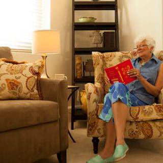 An elderly woman sits comfortably in an armchair, enjoying a book in a cozy living room.
