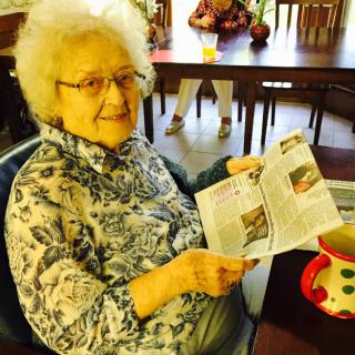 An elderly woman smiling as she reads a newspaper while sitting indoors with a mug on the table.