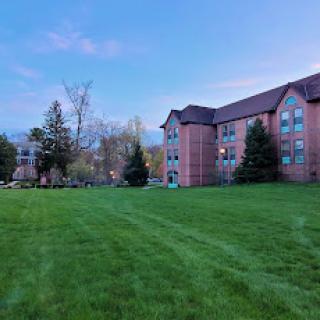 A peaceful view of a brick apartment building surrounded by lush green grass and trees under a clear sky.