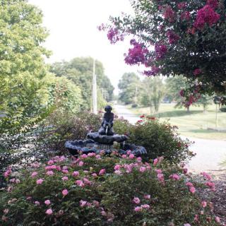 A garden scene with a small statue surrounded by blooming flowers and lush trees.