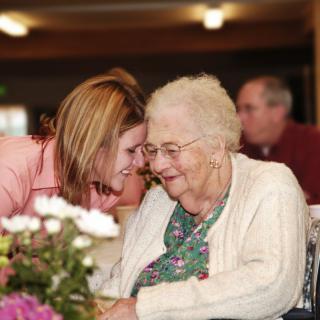 A younger woman whispers affectionately to an elderly woman, surrounded by flowers indoors.