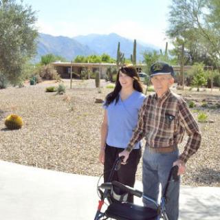 An elderly man with a walker and a young woman walking together in a sunny desert landscape.