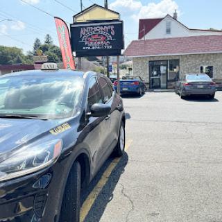 A black taxi is parked outside a restaurant with visible signage in a sunny urban setting.