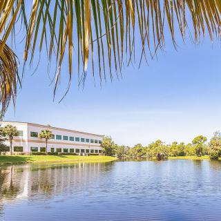 A serene view of a building by a lake surrounded by palm trees under a clear blue sky.