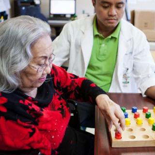 An elderly woman engages in a therapy exercise under the supervision of a healthcare professional.