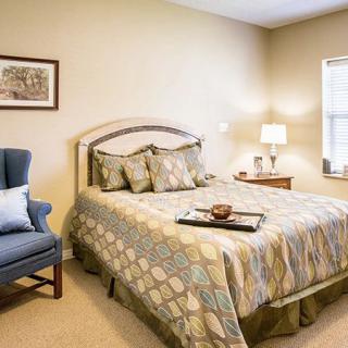 A cozy bedroom with a neatly made bed, an armchair, a bedside lamp, and a window allowing natural light to illuminate the room.