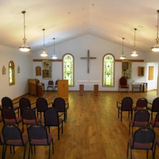 Interior view of a chapel featuring stained glass windows and arranged seating with a cross on the wall.