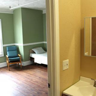 A simple and calm room featuring a bed, chair, and a bathroom area.