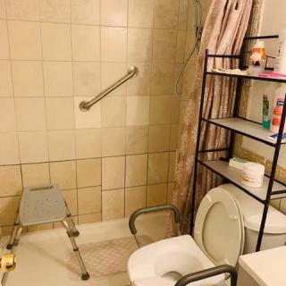 A well-equipped bathroom featuring a toilet with a support rail, a shower area with a bath chair and grab bar, and storage shelves filled with various toiletries.