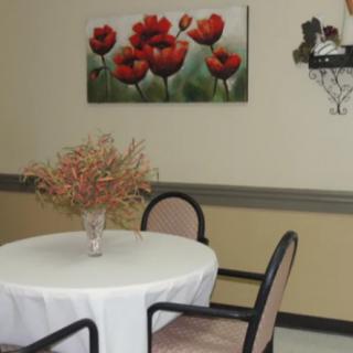 A small round table with a white tablecloth, accompanied by chairs, features a vase with dried flowers. A vivid flower painting decorates the wall.