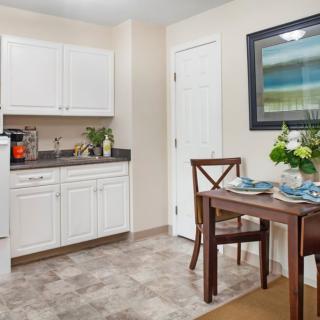 A cozy kitchen and dining area with white cabinets, a small table set for two with flowers, and artwork on the wall.