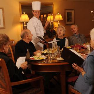 A chef presenting the menu to a group of elderly diners in a cozy restaurant setting.
