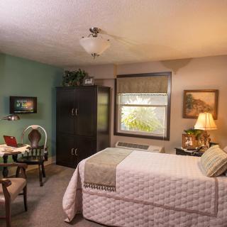 A cozy bedroom with a neatly made bed, a lamp on a side table, and a window letting in natural light.