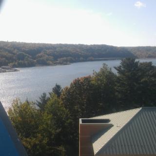 A scenic view of a lake surrounded by hills and trees, with a partial view of a roof in the foreground.