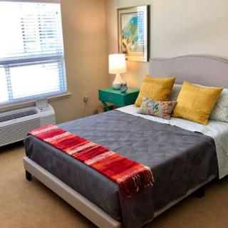 A cozy bedroom with a neatly made bed, featuring colorful pillows and a red-striped throw blanket, next to a teal nightstand and a lamp.
