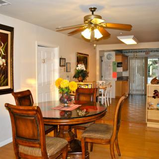 A cozy dining area with a round wooden table and chairs, adorned with a floral centerpiece, leads into an inviting kitchen. A ceiling fan provides light and circulation, complementing the warm tones of the decor.
