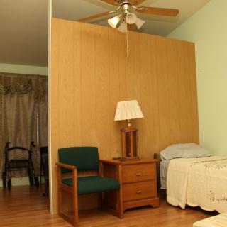 A cozy bedroom with a single bed, nightstand, lamp, and chair, featuring a ceiling fan and wooden flooring.