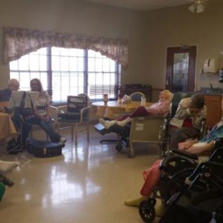 A group of senior citizens sitting in a nursing home common area, some in wheelchairs.