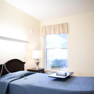 A minimalist bedroom featuring a neatly made bed with a blue cover, a bedside lamp, and a window letting in natural light.