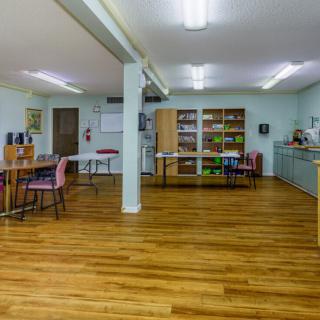 A spacious classroom with wooden floors, tables, chairs, and shelves under bright lighting.