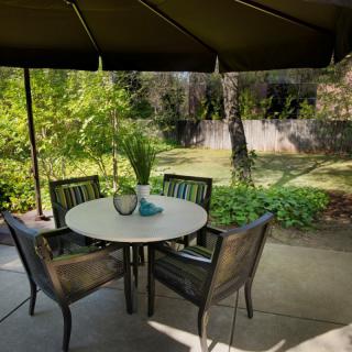 A peaceful outdoor patio with a round table and chairs, shaded by a large umbrella, surrounded by lush greenery.