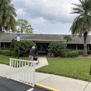 A serene scene featuring a well-maintained building surrounded by palm trees and lush greenery, with an elderly person walking along the pathway.