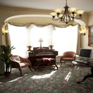 A cozy living room with antique-style furnishings, featuring a chandelier, a computer desk, and large windows with curtains.