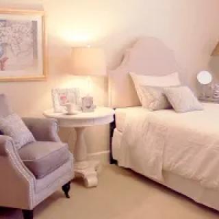 A cozy and elegant bedroom with a soft color palette, featuring a bed, armchair, lamp, and wall art.