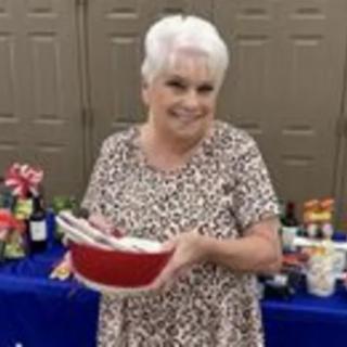 A woman smiling and holding a bowl at an indoor gathering with food in the background.